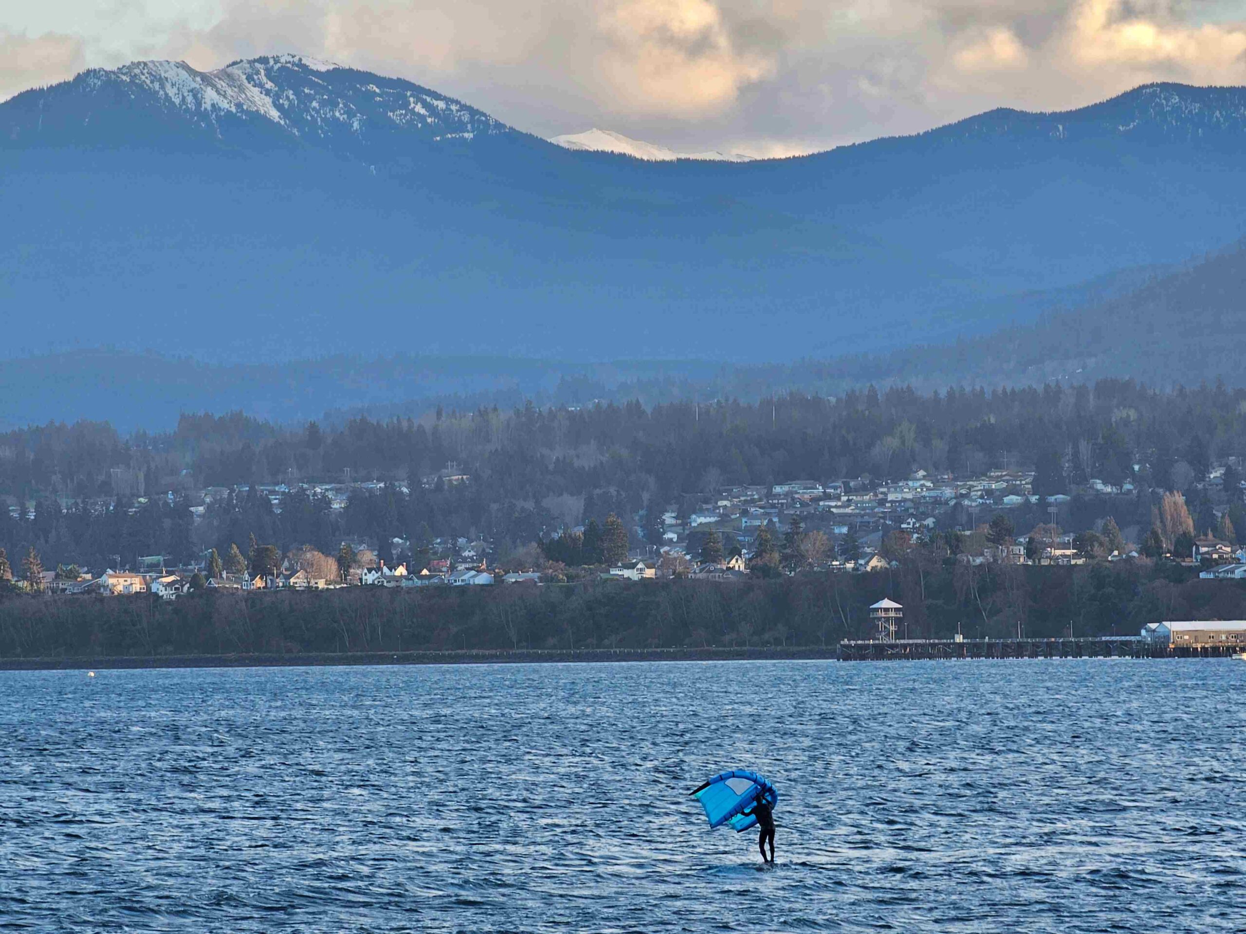 Photograph of Port Angeles Harbor, looking south from Ediz Hook with Olympic Mountains in background, blue water and kiteboarder with blue kite